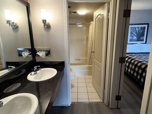 A hotel bathroom with two sinks, a shower, and a bed visible through an open door.