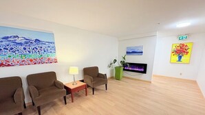 A spacious room with a wooden floor, two brown armchairs, a small wooden table with a lamp, a green plant, a flat-screen TV, and two paintings on the walls.