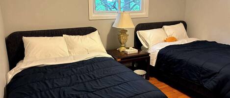 Habitación con dos camas, cada una con ropa de cama azul oscuro y almohadas blancas. Hay una mesita de noche con una lámpara entre las camas. Se ve una ventana con persianas que muestra follaje en el exterior.