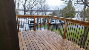 A wooden deck with a metal railing overlooking a body of water, a grassy area, and a parked vehicle.