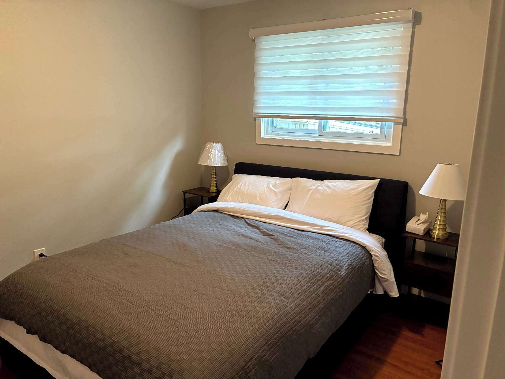 Una cama bien hecha con almohadas blancas y una manta gris, una lámpara de noche y una ventana con persianas.