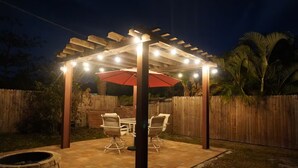 En uteplats med en pergola, ljusslingor, ett bord, stolar och en eldstad.