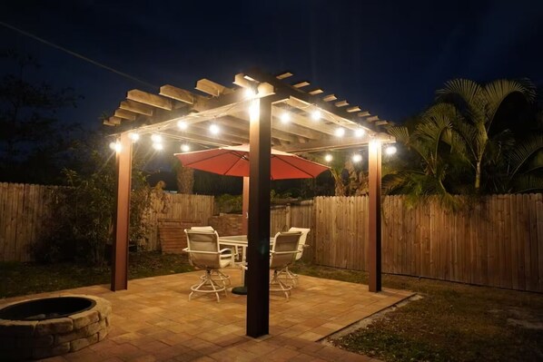 Un patio al aire libre con pérgola, luces colgantes, una mesa, sillas y un brasero.