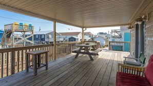 A wooden deck with picnic tables and a wicker chair, overlooking a residential area with houses and a dock.