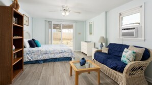A room with a bed, a wicker sofa, a wooden bookshelf, a glass coffee table, and a ceiling fan.