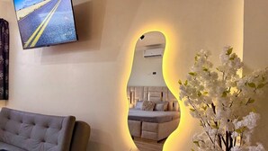 A living room with a sofa, a mirror with a yellow light, and a wall-mounted TV displaying a road scene. - Master Suite Plus Ocean View Balcony (San Cristóbal)
