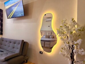 A living room with a sofa, a mirror with a yellow light, and a wall-mounted TV displaying a road scene.