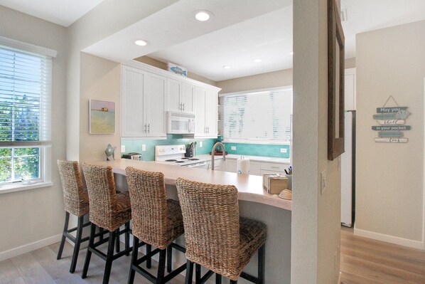 A kitchen with white cabinets, a central island with bar stools, a microwave, and a window with blinds.