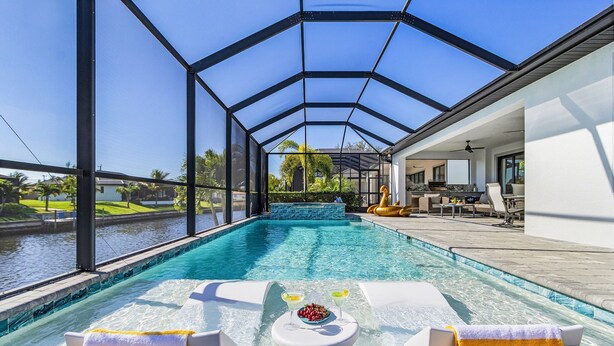 A pool area with a glass-enclosed structure, lounge chairs, and a table with drinks and fruit.