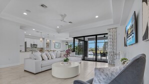 A modern living room with a grey armchair, white sofa, and a coffee table. There is a kitchen area with white cabinets and a dining area with a glass table.