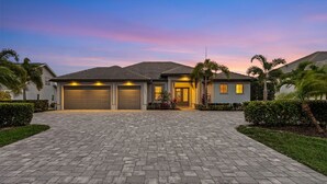 A modern two-story house with a paved driveway, garage doors, and a well-maintained lawn with palm trees.