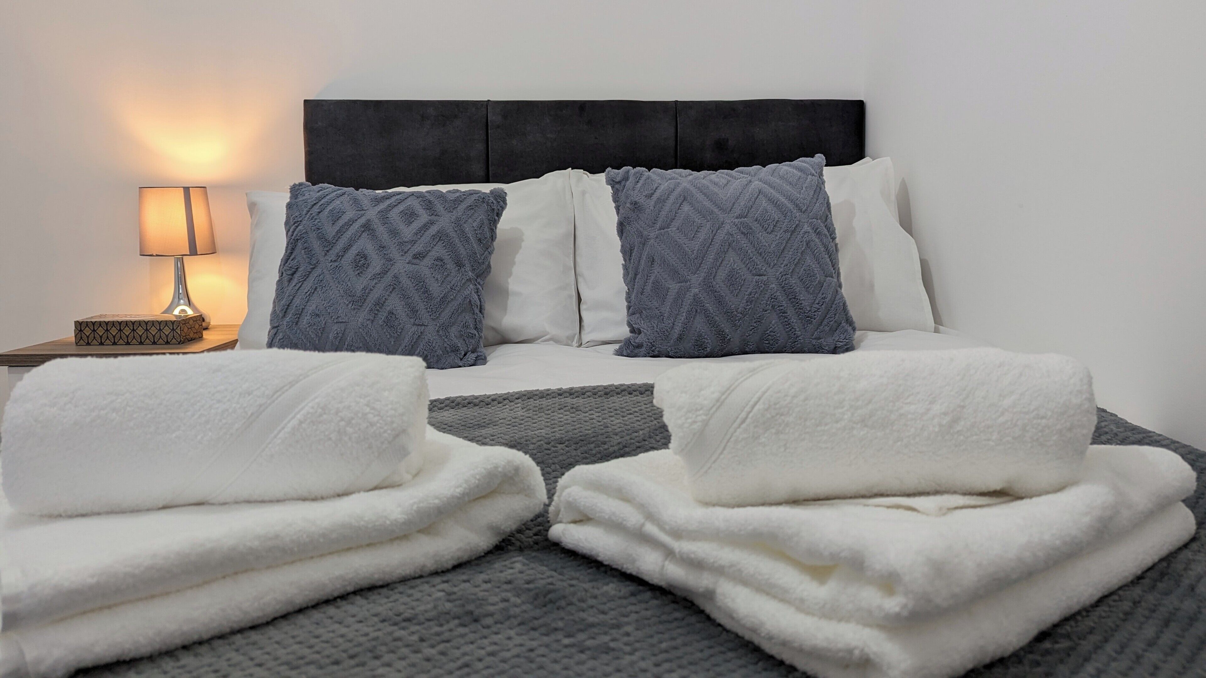 Een net opgemaakt bed met witte en grijze dekens, een bedlampje en een kussen met een patroon.