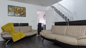 Un salon avec deux canapés beige, un plaid jaune, un escalier et une horloge au mur.
