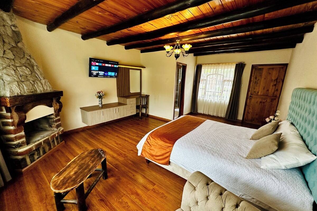 Ein Schlafzimmer mit Kamin aus Stein, Holzbalken an der Decke, einem Bett mit Quilt, einer Bank und einem Fernseher an der Wand.