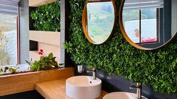 Un baño moderno con dos lavamanos redondos, un mueble de tocador de madera, una pared verde con plantas y un espejo grande.