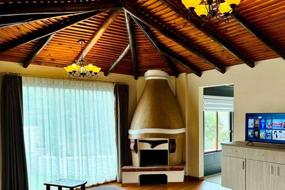 Ein geräumiges Zimmer mit einem großen Bett, einer Holzbalkendecke, einem großen Fenster mit Vorhängen, einem Fernseher und einem Schrank.