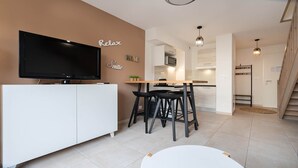 A modern living room with a flat-screen TV, a white cabinet, and a dining area with a wooden table and black bar stools. - Villa duplex 6 pers near the beach (Sérignan)