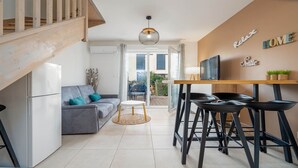 A modern living room with a grey sofa, a small round table, and black bar stools. There is a TV on a wooden table, and a staircase on the left. - Villa duplex 6 pers near the beach (Sérignan)