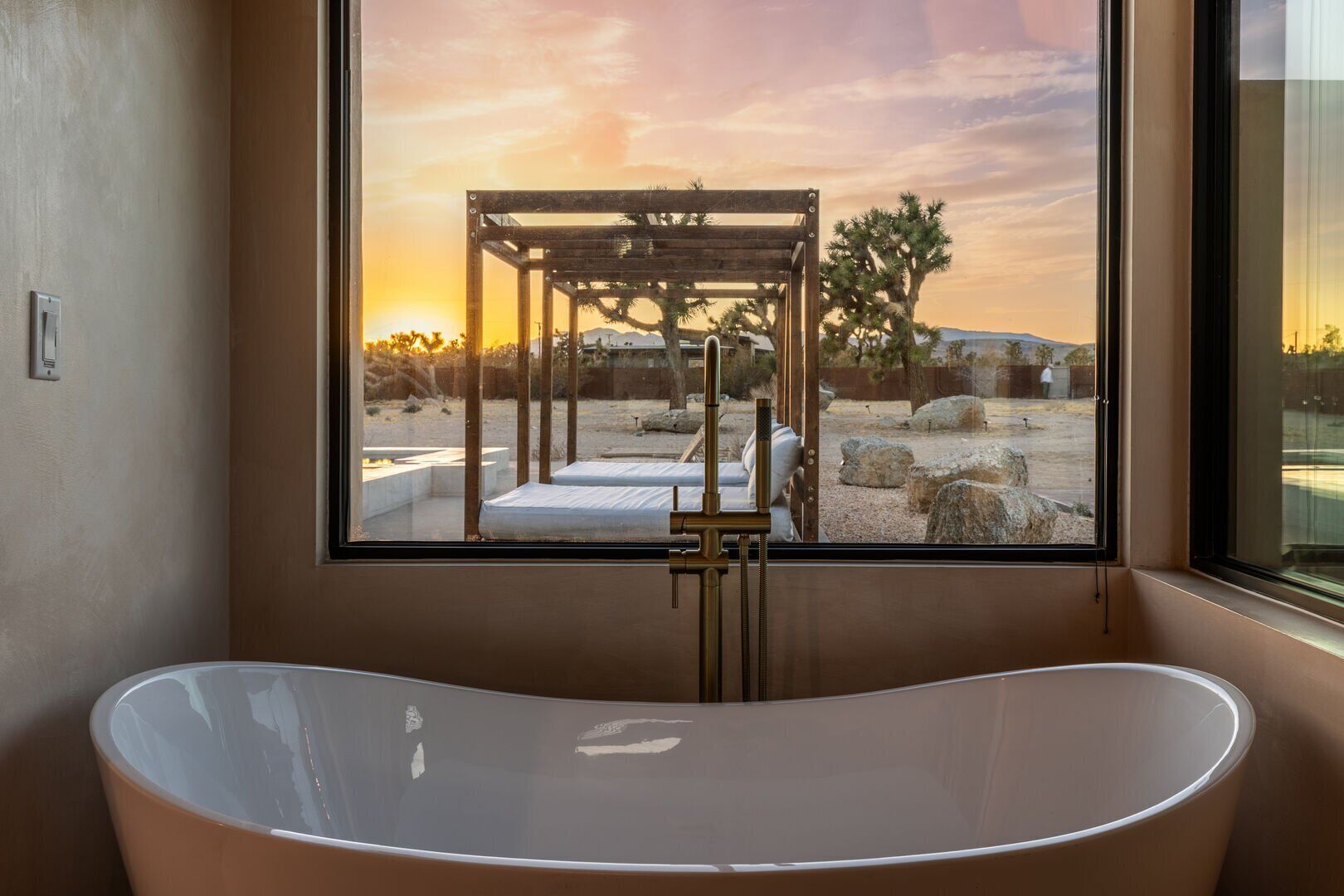 A modern bathroom with a freestanding bathtub, a large window with a view of a desert landscape, and a wooden structure in the window.