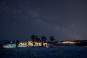 Eine Wüstenlandschaft bei Nacht mit einem Haus, Palmen und einem Sternenhimmel.