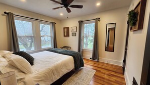 Ein Schlafzimmer mit einem großen Bett, zwei mit Vorhängen versehenen Fenstern, einem Deckenventilator und einem Holzboden.