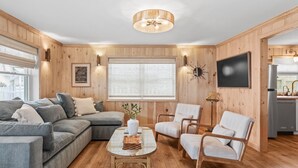 Ein gemütliches Wohnzimmer mit einer Eckcouch, einem Couchtisch und zwei Sesseln. Ein großes Fenster sorgt für natürliches Licht, und ein Flachbildfernseher ist an der Wand montiert.