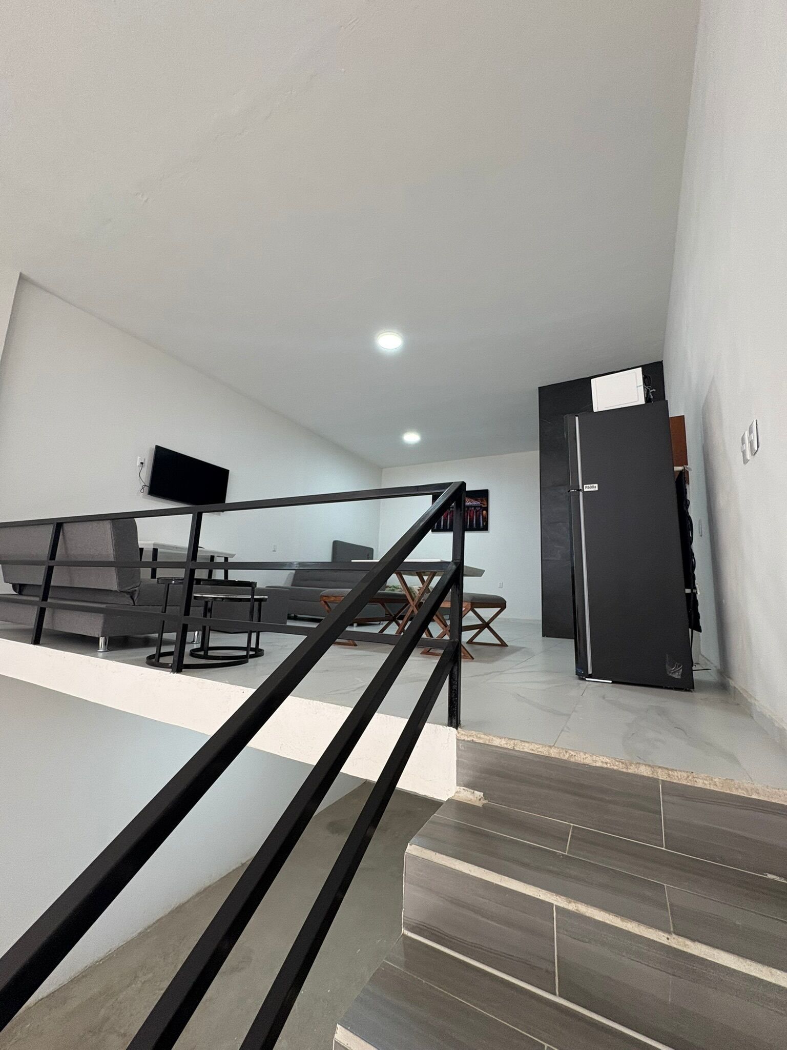 A modern living room with a sofa set, a flat-screen TV mounted on the wall, and a black refrigerator.