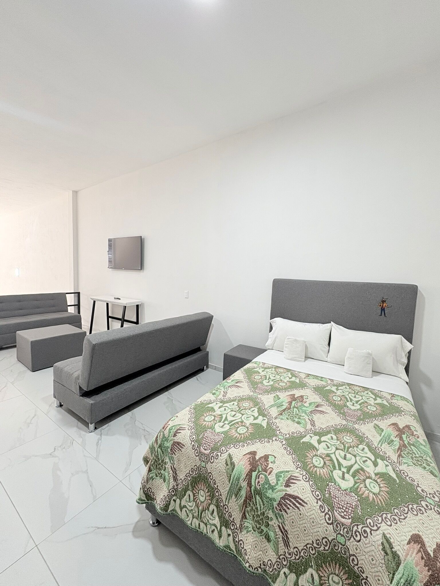 A modern bedroom with a large bed, grey sofas, a small table, and a TV on the wall.