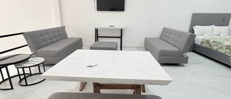 Una sala moderna con una mesa blanca grande, sofás grises, una mesita redonda pequeña y una cama con cabecera gris.
