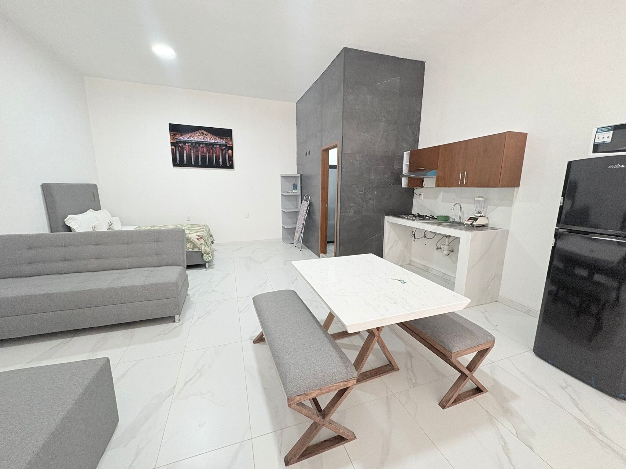 Un espacio habitable funcional con sofá, mesa de comedor con bancos, zona de cocina con armarios y electrodomésticos, y una obra de arte colgada en la pared.