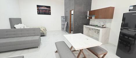 Un espacio habitable funcional con sofá, mesa de comedor con bancos, zona de cocina con armarios y electrodomésticos, y una obra de arte colgada en la pared.