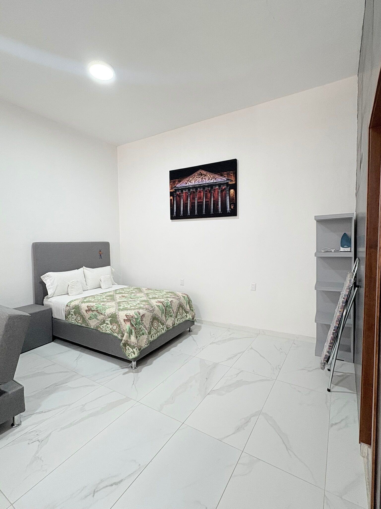 Ein Schlafzimmer mit einem Bett, einer grauen Kopfteil, einem Marmorboden, einem Wandbild und einem kleinen Regal mit einem Bügeleisen.
