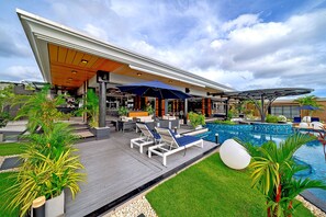 Un área de piscina moderna al aire libre con sillones de descanso y sombrillas, rodeada de vegetación y una zona de bar.