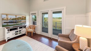 A living room with a television, a wicker chair, a sofa, and a lamp. There is a sliding glass door leading to a balcony with a view of a bridge and water.