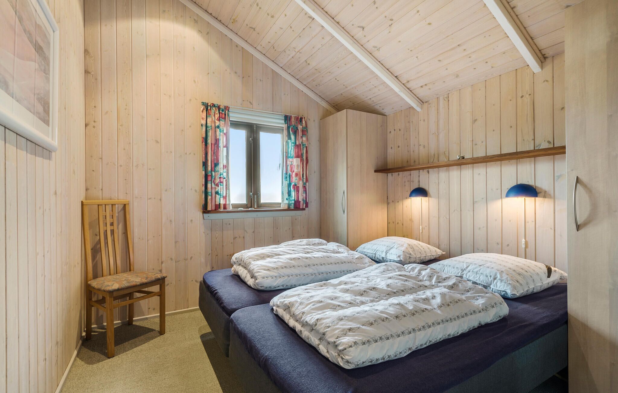 Quarto com paredes e teto de madeira, duas camas com roupa de cama floral, uma cadeira, janela com cortinas e luminárias fixadas na parede.