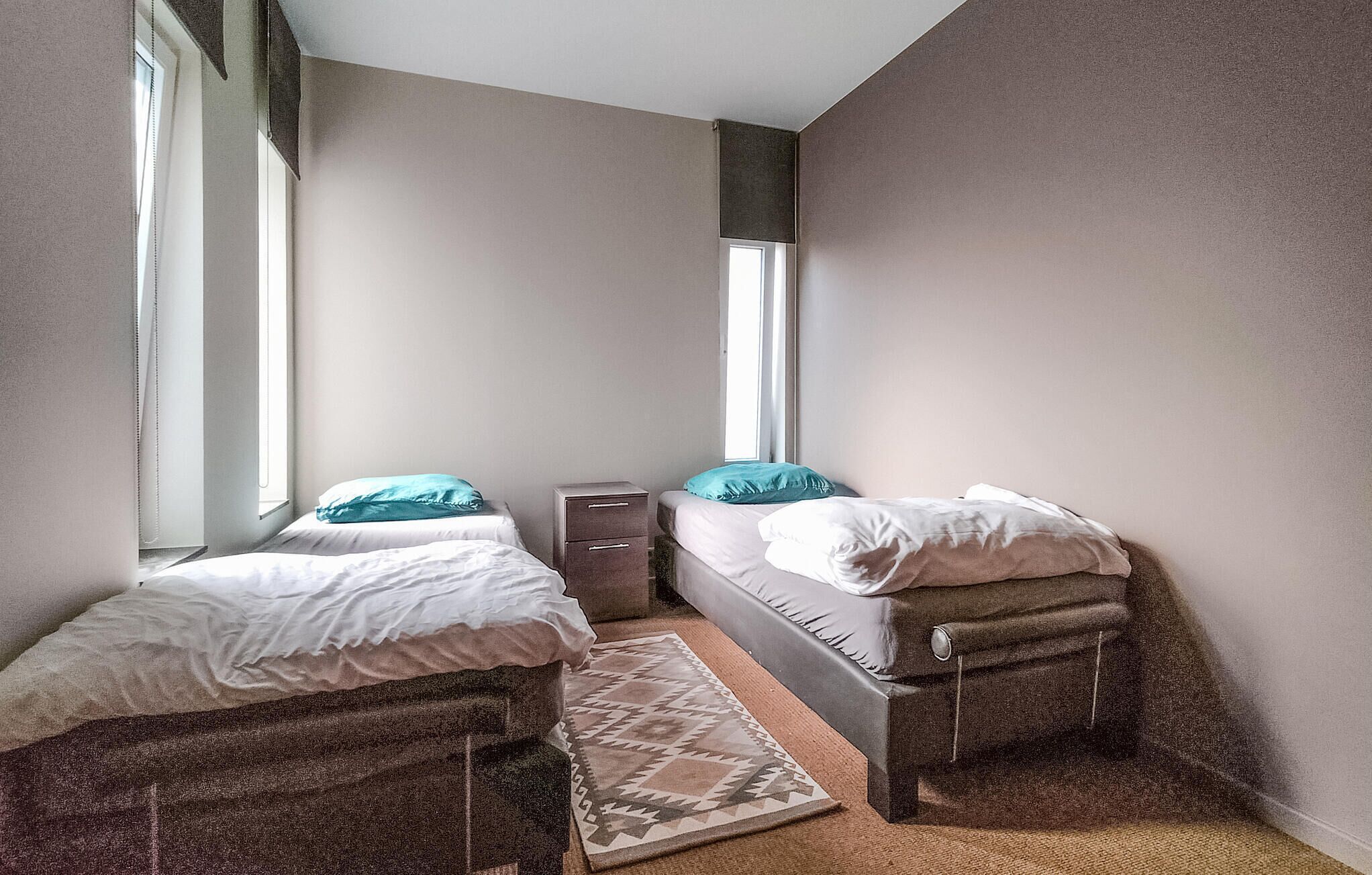 A small, compact room with two single beds, a small bedside table, and a window providing natural light.