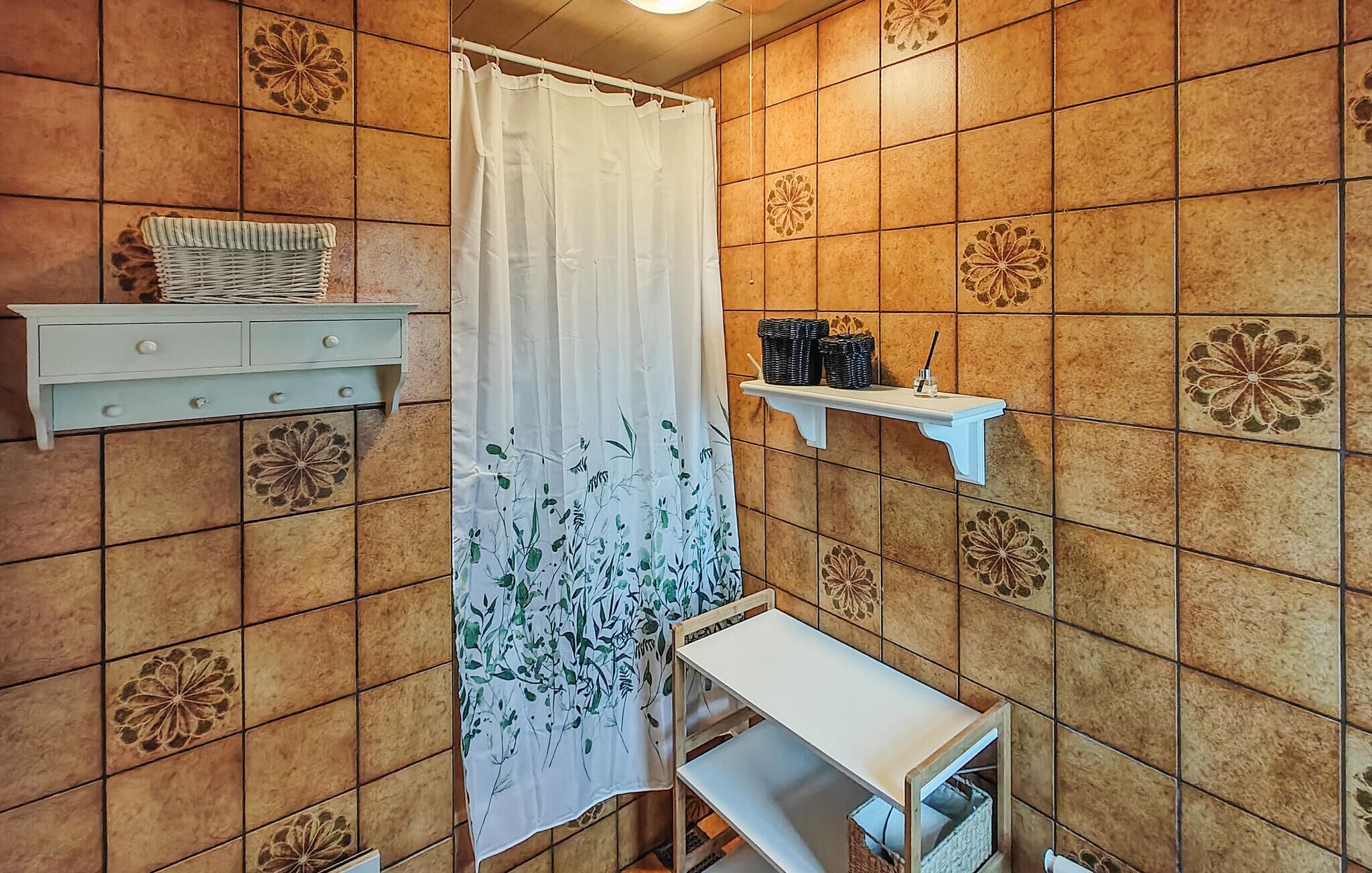 A bathroom with a shower curtain, a small shelf with toiletries, and a tiled wall with floral patterns.