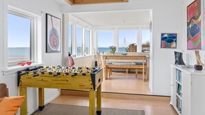 Una habitación con mesa de fútbol, zona de comedor con mesa y sillas, y ventanas amplias con vista al mar.