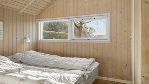 A bedroom with wooden walls, a bed with white bedding, a window with a view of trees and a hedge, and a wall-mounted lamp.