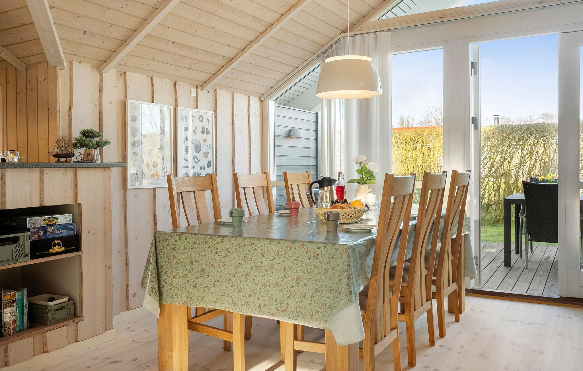 Un coin repas avec une table dressée pour six personnes, des chaises en bois, un luminaire suspendu et une vue sur l’extérieur grâce à de grandes fenêtres.