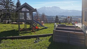 Un parque infantil con tobogán y columpios, una terraza de madera y vistas a montañas al fondo.