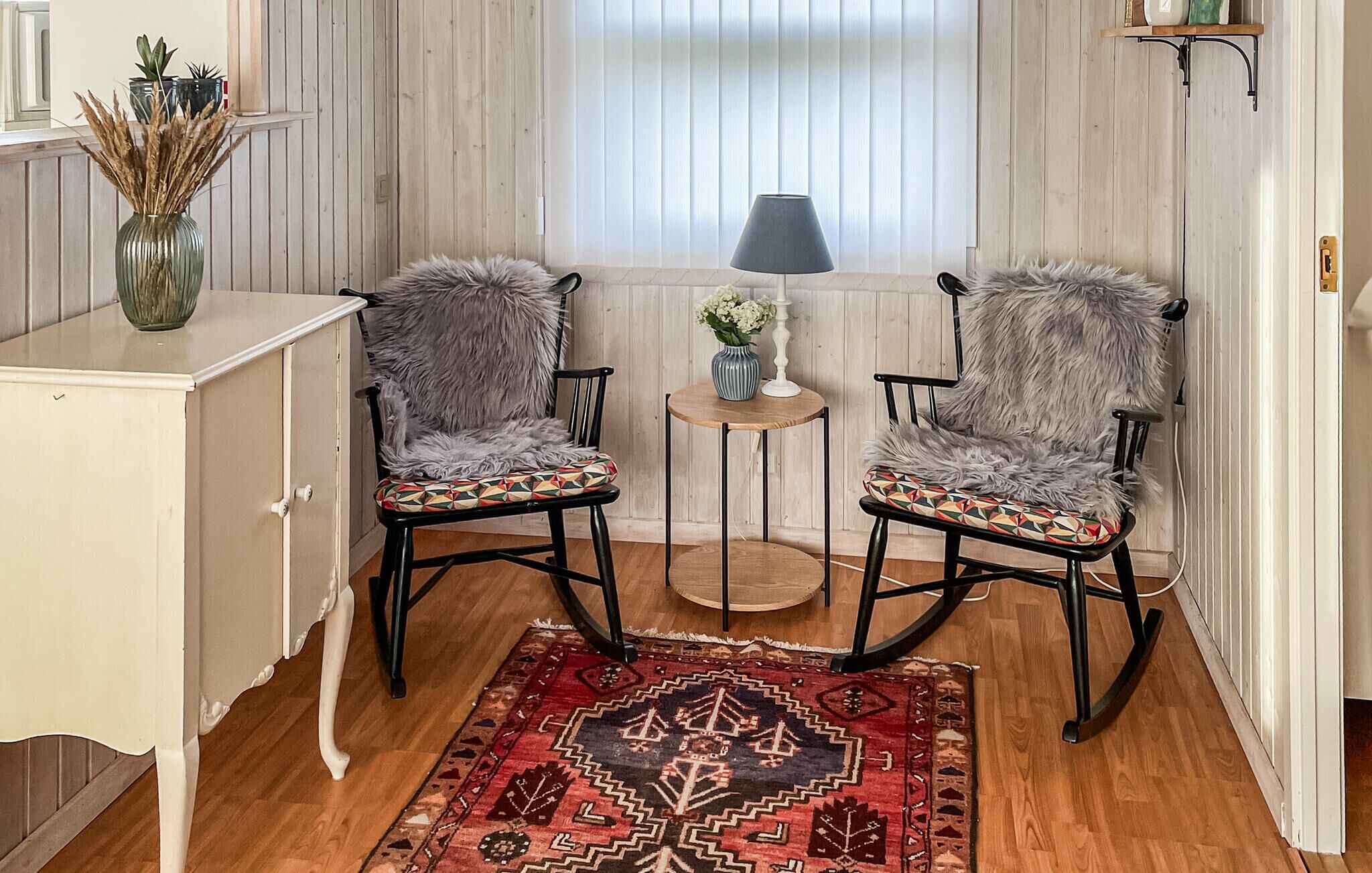 A room with two rocking chairs, a small round table with a vase and flowers, and a patterned rug on the wooden floor.