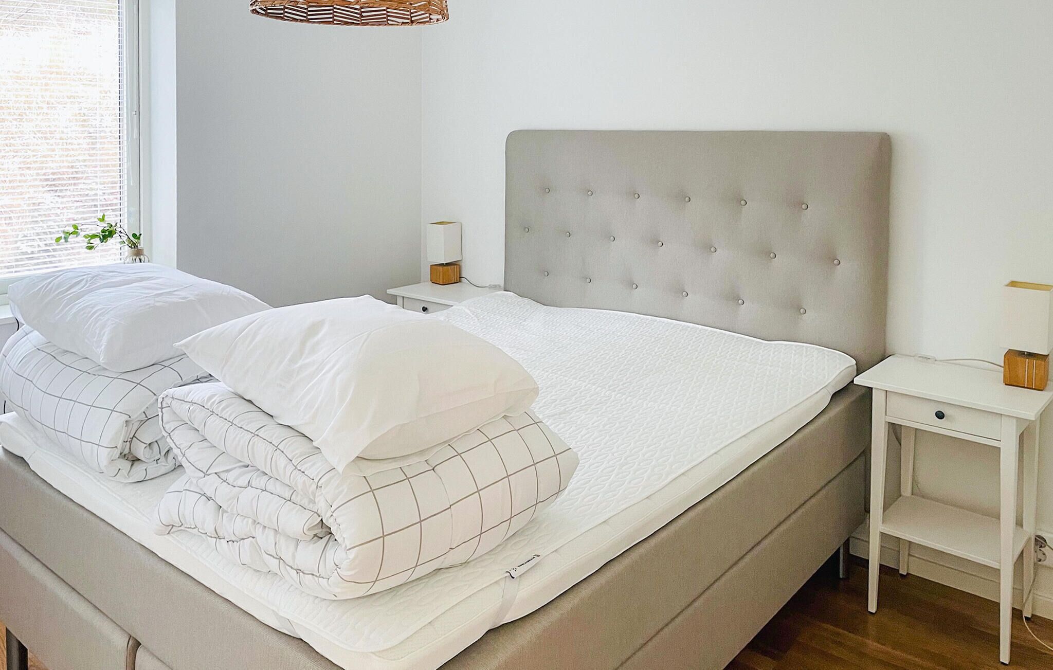A neatly made bed with white bedding and a grey headboard, a bedside table with a lamp, and a small plant on the windowsill.