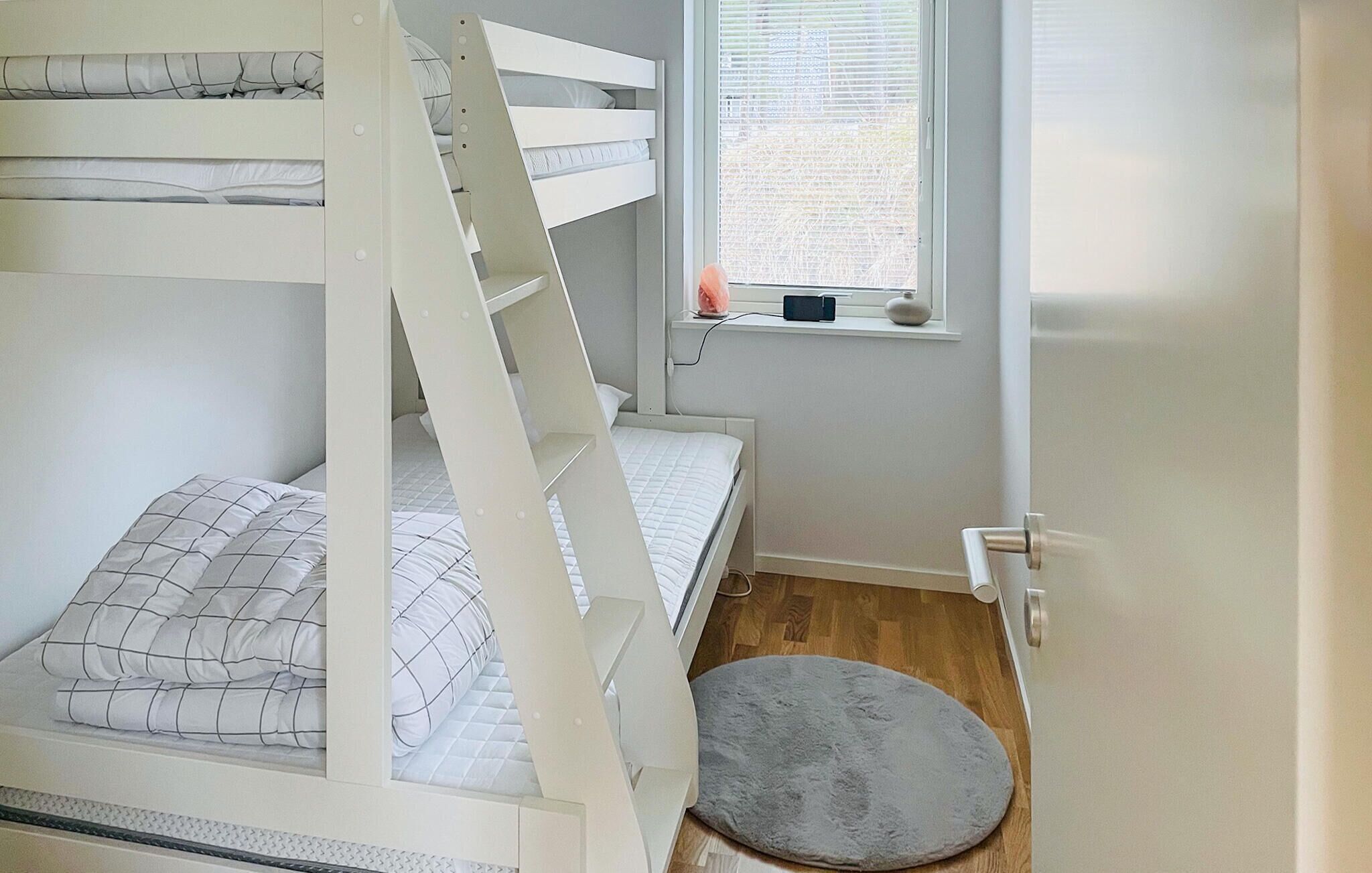 En våningssäng med ett fönster ovanför, en liten matta och en dörr till höger.