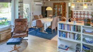 A cozy living room with a leather armchair, a dining table with chairs, a bookshelf, and a kitchen area with a wooden cabinet and a hanging light fixture. - 4 bedroom amazing home in Hurup Thy (Hurup Thy)