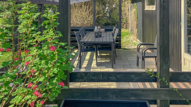 An outdoor patio with a wooden table and chairs, surrounded by a wooden pergola and a garden with green plants and red flowers. - 4 bedroom amazing home in Hurup Thy (Hurup Thy)