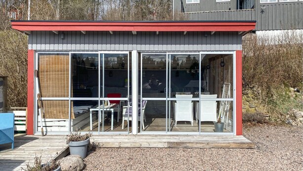 A modern, single-story building with a red roof and large glass windows. It features a wooden deck with outdoor furniture and potted plants. - 1 bedroom stunning home in Mullsjö (Mullsjö)