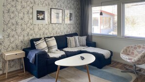 A living room with a dark blue sofa, a white coffee table, and a patterned armchair. There are framed pictures on the wall and a window with a view of buildings outside. - 1 bedroom stunning home in Mullsjö (Mullsjö)