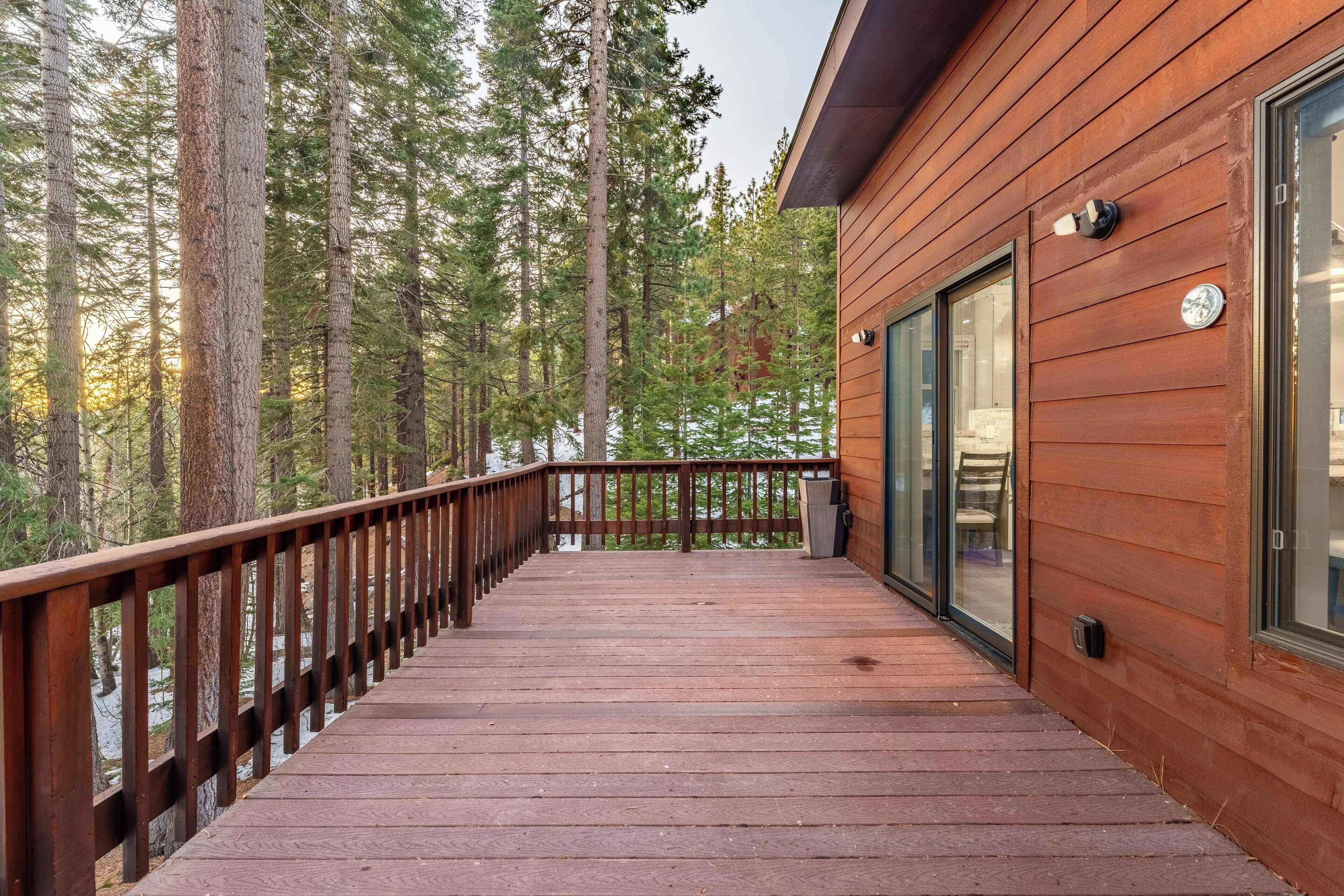Une terrasse en bois, dotée d’une rambarde et entourée de grands arbres, mène à un bâtiment aux larges fenêtres.
