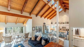 A spacious living area with a high ceiling and wooden beams, a cozy seating area with a dark blue sofa and wooden coffee table, and a view of the kitchen and dining area through large windows. - Views + Mins to Skiing, Lake, & Incline Village Incline Serenity by Avantstay (Incline Village)
